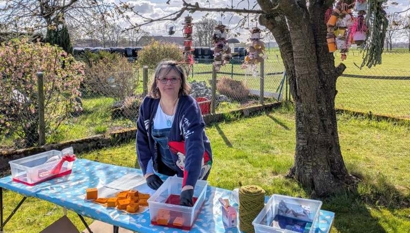 So werden die bunten Balsaholzklötze und buntes Papier hergestellt. Susanne steht draußen und färbt mit lebensmittelechten Farben Balsaholzklötze. Im Baum hängt bereits fertiges Becherglück-Spielzeug.